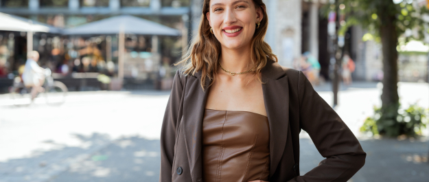 Woman in a brown top with a blazer, outside on the street.