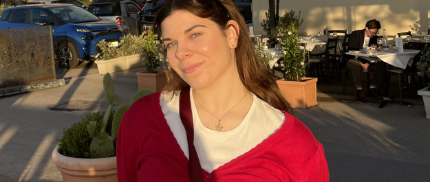 Young woman with long brown hair wears a red sweater, posing in the sun.
