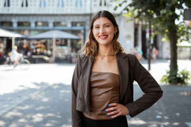 Woman in a brown top with a blazer, outside on the street.