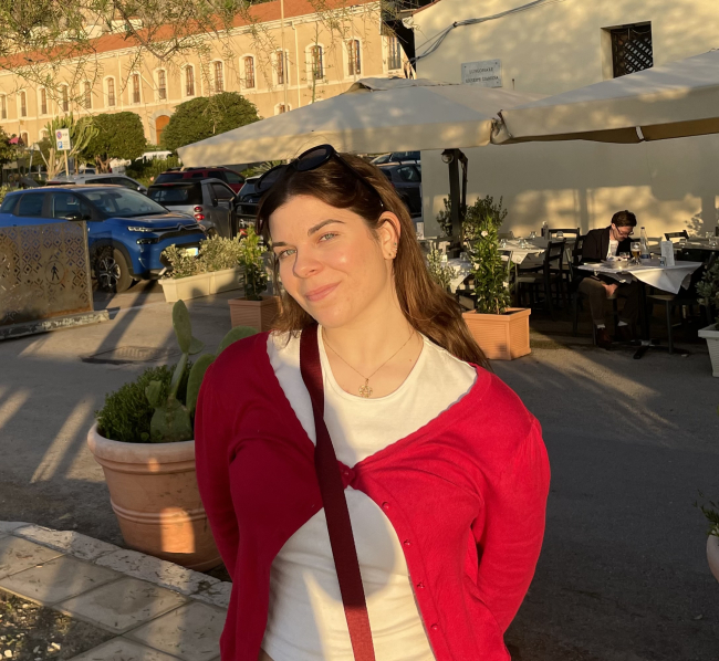 Young woman with long brown hair wears a red sweater, posing in the sun.