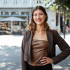 Woman in a brown top with a blazer, outside on the street.