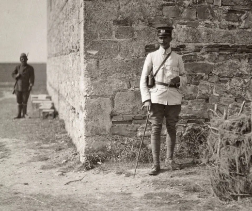 Major Roelfsema standing with cane and binoculars beside a stone wall at the location in Durrës, Albania, where Major Thomson was fatally shot during the 1914 peace mission. An armed soldier stands guard in the background.