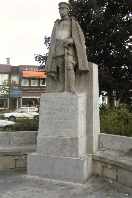 Stone statue of Major L.W.J.K. Thomson in military uniform and cape, standing atop a pedestal at Thomsonplein in The Hague, flanked by a semicircular stone bench and set against a backdrop of local shops.