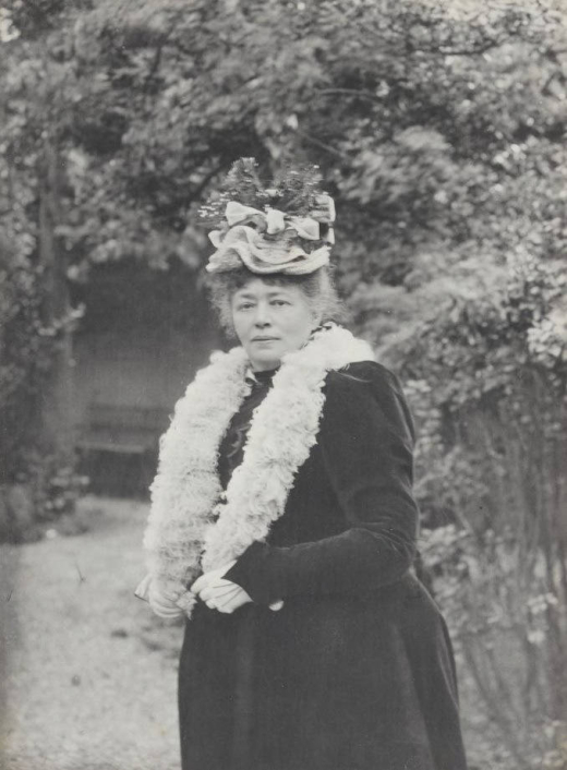 A woman with a unique hat and fur collar poses in a green garden.