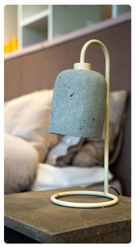 A modern lamp with a concrete shade and yellow base, on a table with cushions in the background.
